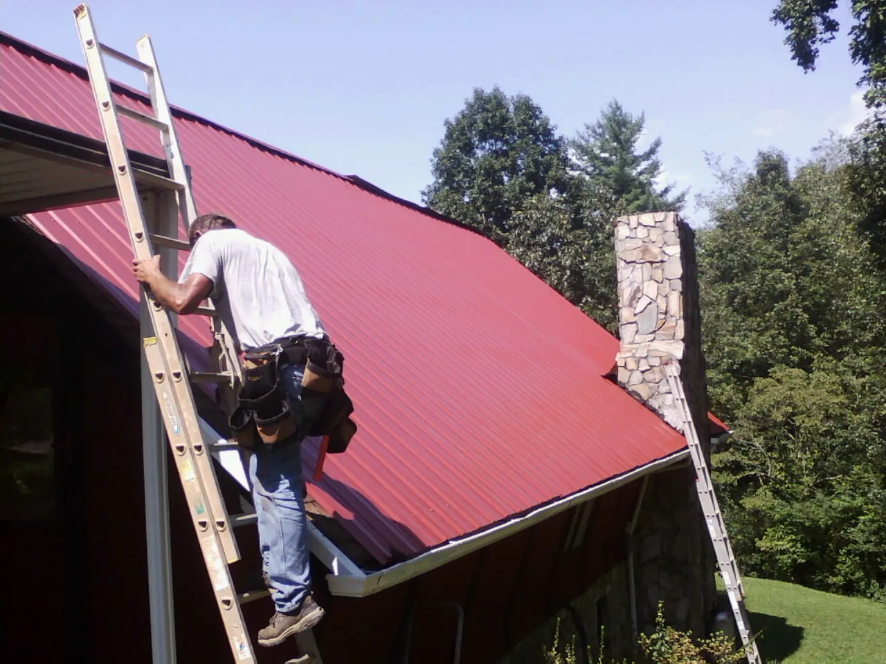 Roofer on a ladder beside a red metal roof and stone chimney — Soffit & Fascia Repair by Roofing Brothers in Port Penn
